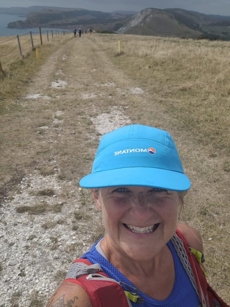 Smiling runner in a blue hat and red running backpack with backdrop of cliffs and coastline